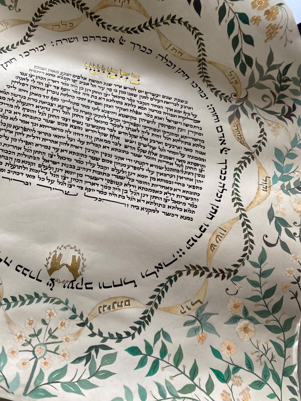 La parte inferiore accosta il piccolo motivo delle mani alla vegetazione esterna; le foglie verde petrolio e i fiori chiari bilanciano il fitto testo nero.
