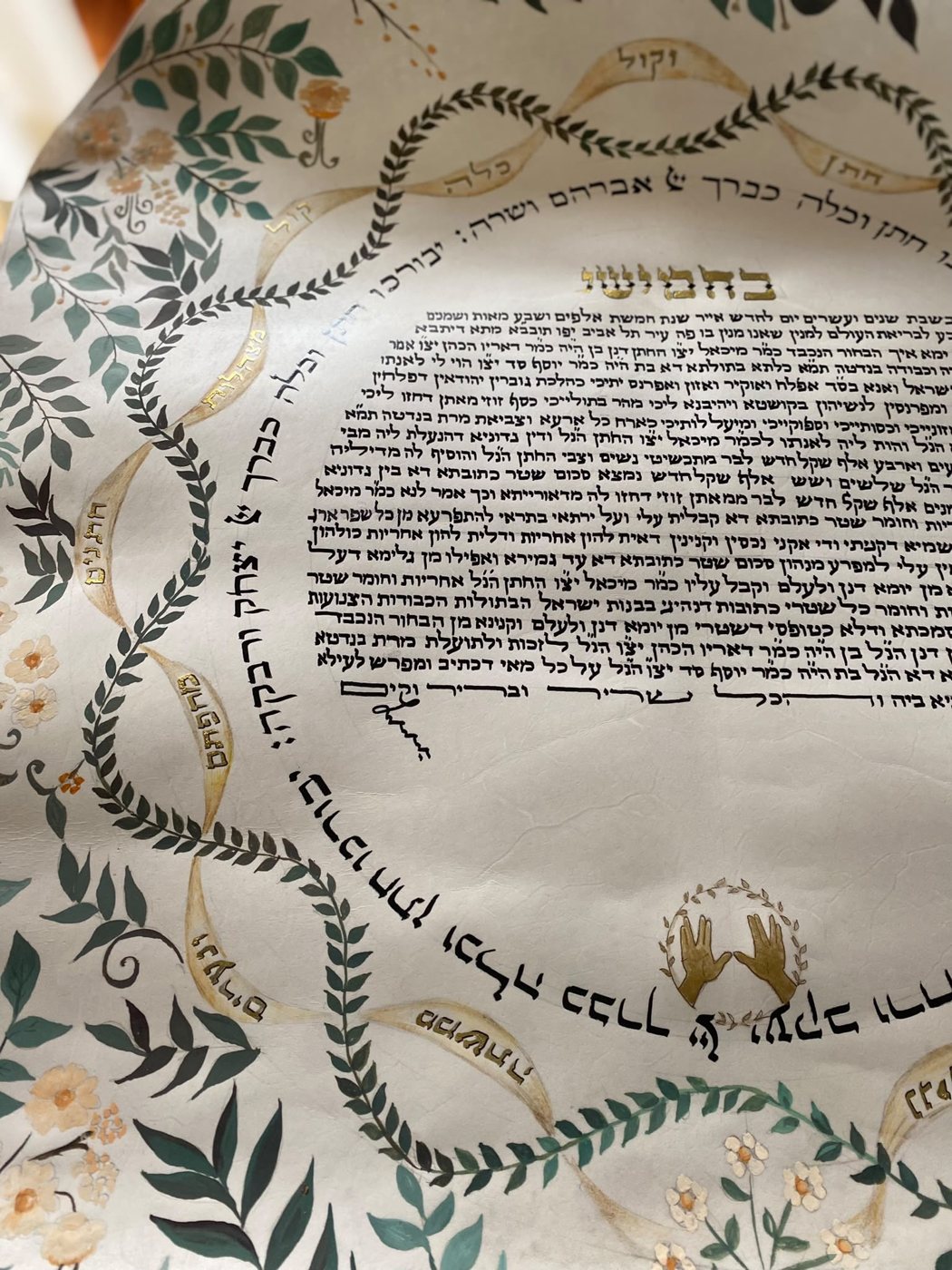 Lo sguardo si concentra sulla linea calligrafica interna e sul motivo delle mani, incorniciati da nastri chiari, foglie scure e minute presenze floreali.